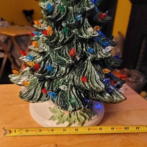 May include: A ceramic Christmas tree with a green glaze and multi-colored lights. The tree is 11 inches tall and has a white base.