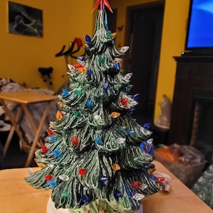 May include: A ceramic Christmas tree with a green glaze and multi-colored lights. The tree is decorated with a red star on top and has a white base.