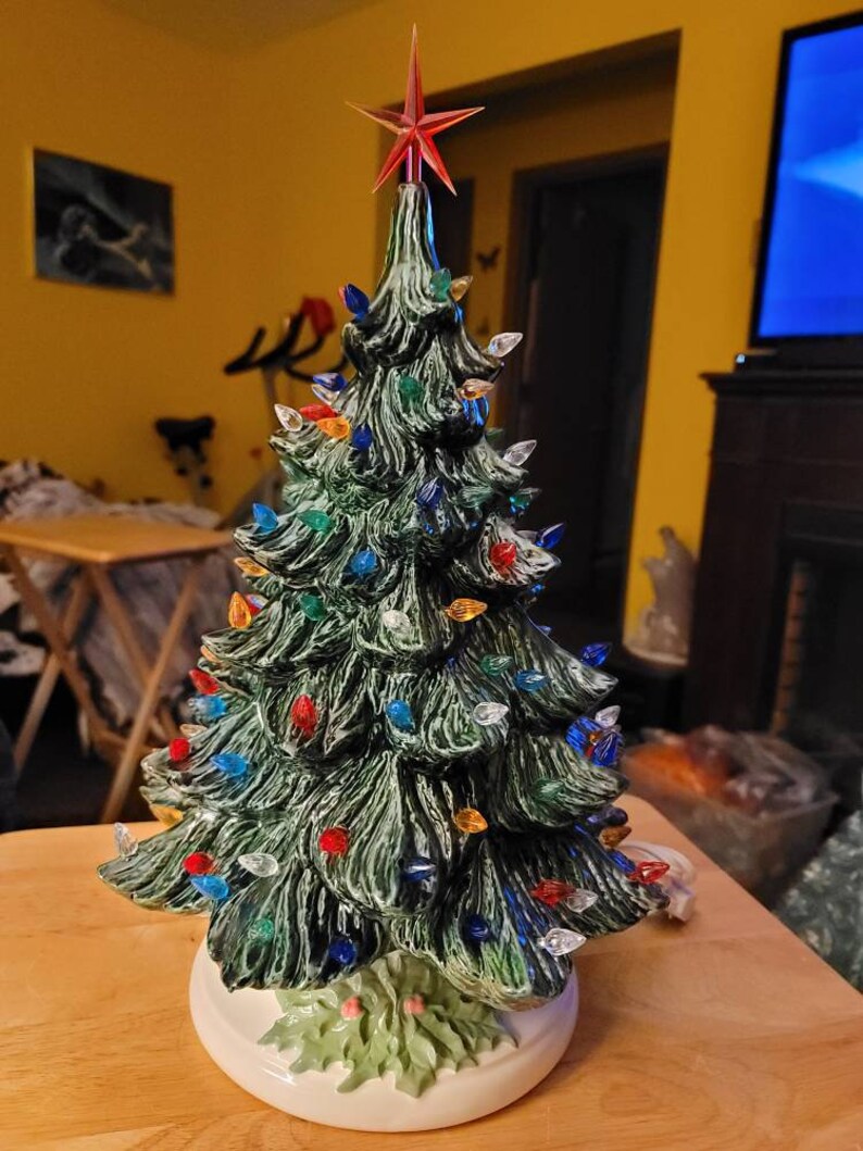 May include: A ceramic Christmas tree with a green glaze and multi-colored lights. The tree is decorated with a red star on top and has a white base.