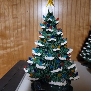 May include: A ceramic Christmas tree with a green glaze and white snow-like accents. The tree is decorated with colorful lights and a gold star on top.