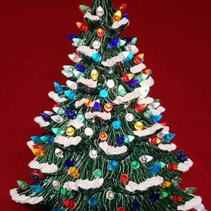 Puede incluir: Un árbol de Navidad de cerámica con una base verde y nieve artificial blanca. El árbol está decorado con luces de colores y una estrella en la parte superior.