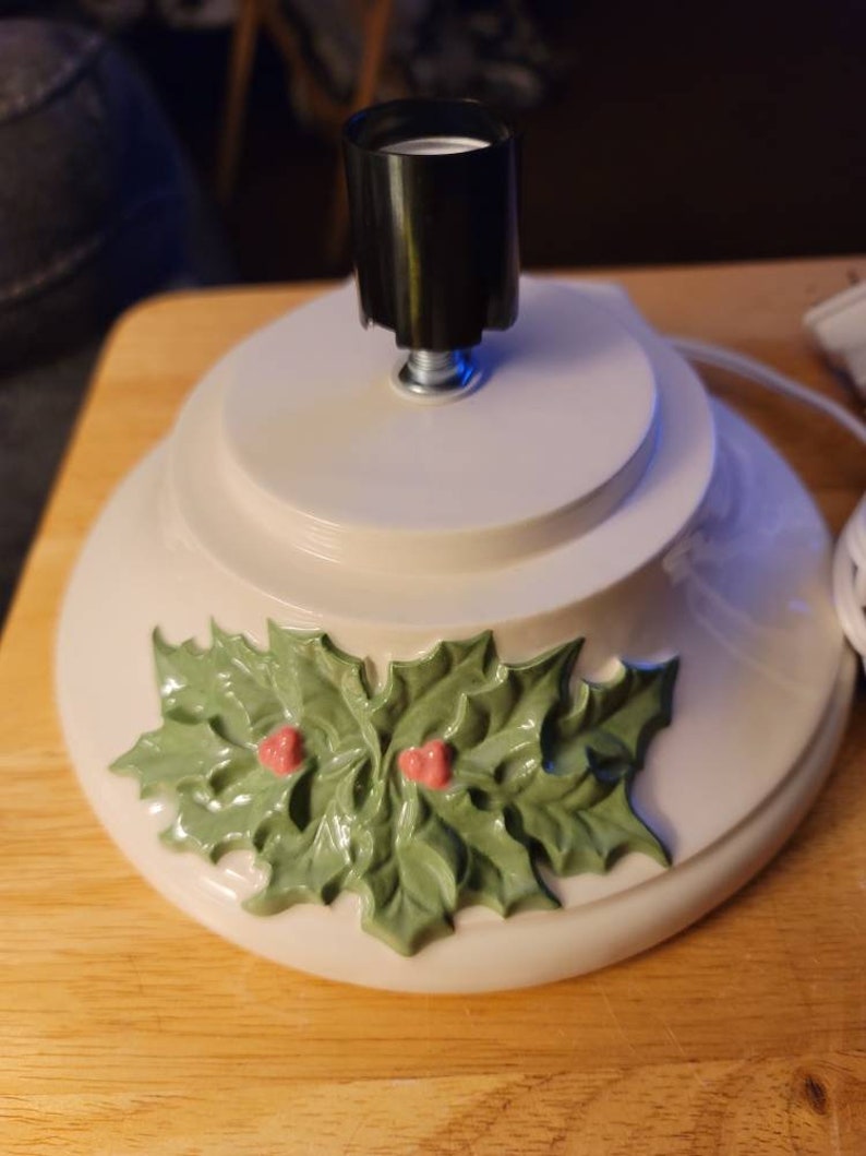 May include: White ceramic Christmas tree stand with a holly leaf design and a black light socket.