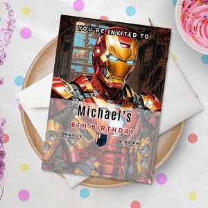 May include: An invitation card featuring Iron Man, with the text "YOU'RE INVITED TO Michael's 6TH BIRTHDAY MAY 16 6:00 PM". The card is on a wooden plate with a white envelope and a cupcake with pink frosting.