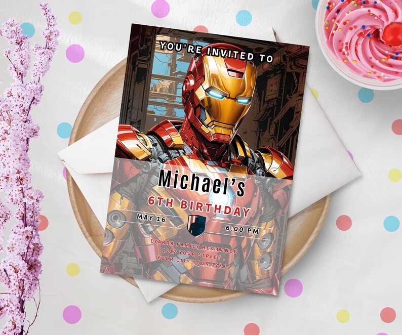 May include: An invitation card featuring Iron Man, with the text "YOU'RE INVITED TO Michael's 6TH BIRTHDAY MAY 16 6:00 PM". The card is on a wooden plate with a white envelope and a cupcake with pink frosting.