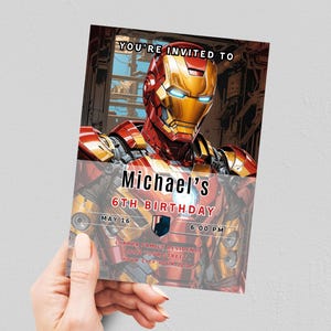 May include: A birthday invitation featuring Iron Man. The invitation has a red and gold color scheme with the text "YOU'RE INVITED TO Michael's 6TH BIRTHDAY MAY 16 6:00 PM". The invitation is held by a person.