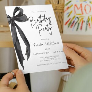 May include: A white birthday party invitation held by a person. The invitation features a large black bow illustration and elegant script text. The text includes the name "Emilia William", date, time, and location.