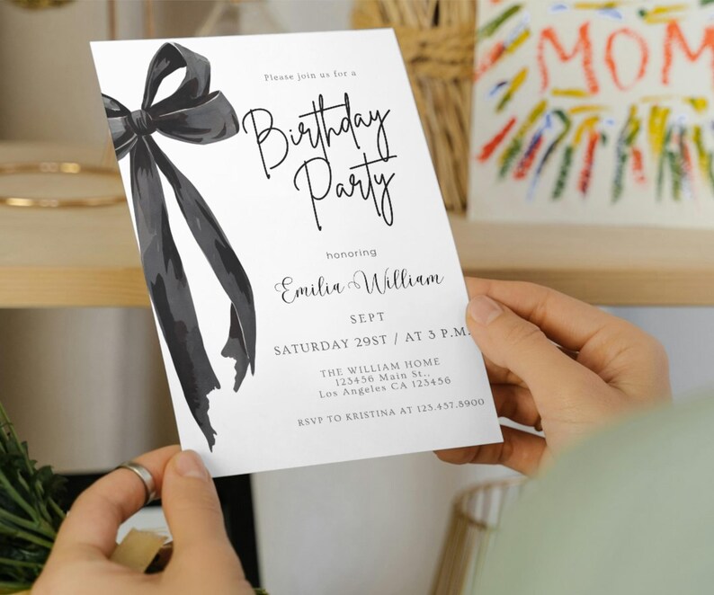 May include: A white birthday party invitation held by a person. The invitation features a large black bow illustration and elegant script text. The text includes the name "Emilia William", date, time, and location.