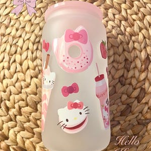 May include: A pink frosted glass featuring Hello Kitty designs. The illustrations include a donut, strawberry, and bows. A drink with a straw and the words "Hello Kitty" in cursive are also visible.