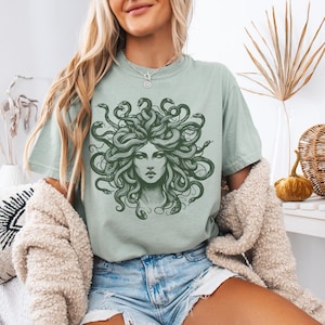 May include: Sage green t-shirt featuring a detailed illustration of Medusa with snakes for hair. The graphic is in a dark green color. The shirt is a casual style, suitable for everyday wear.
