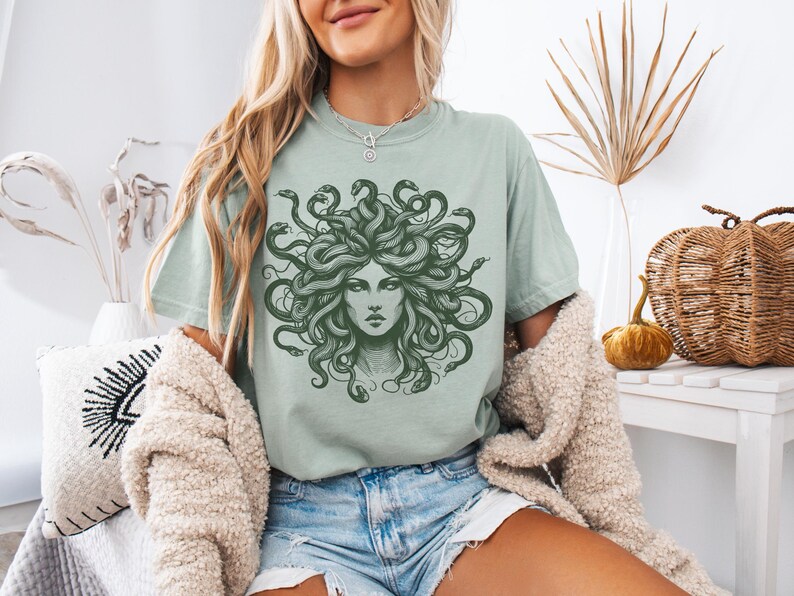 May include: Sage green t-shirt featuring a detailed illustration of Medusa with snakes for hair. The graphic is in a dark green color. The shirt is a casual style, suitable for everyday wear.
