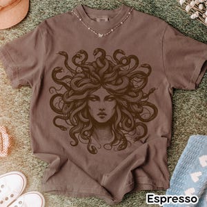 May include: Espresso-colored t-shirt featuring a detailed illustration of Medusa with snakes for hair. The graphic is in a darker brown shade. The shirt has short sleeves and a crew neck. The word "Espresso" is printed at the bottom of the shirt.