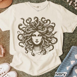 May include: Ivory-colored t-shirt featuring a detailed illustration of Medusa's face with snakes for hair. The graphic is in shades of brown and black. The shirt has a crew neck and short sleeves. The word "Ivory" is printed at the bottom right.