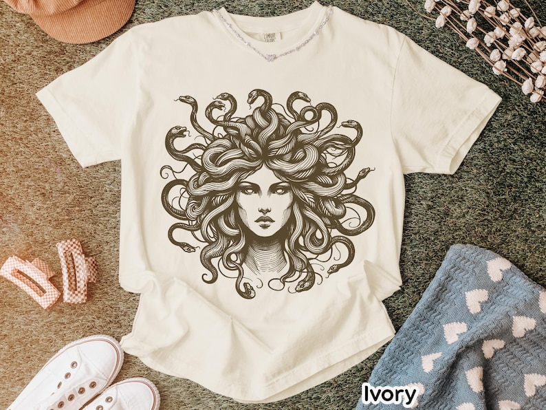 May include: Ivory-colored t-shirt featuring a detailed illustration of Medusa's face with snakes for hair. The graphic is in shades of brown and black. The shirt has a crew neck and short sleeves. The word "Ivory" is printed at the bottom right.
