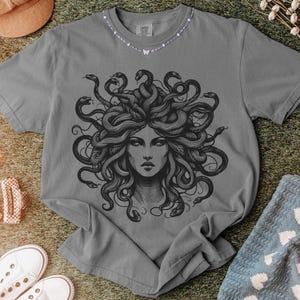 May include: Gray t-shirt featuring a detailed black illustration of Medusa with snakes for hair. The graphic is centered on the shirt. The shirt is displayed on a textured surface with other items.