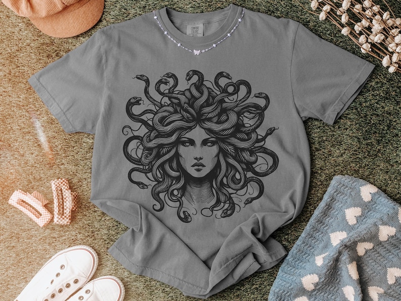 May include: Gray t-shirt featuring a detailed black illustration of Medusa with snakes for hair. The graphic is centered on the shirt. The shirt is displayed on a textured surface with other items.