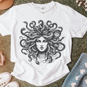 May include: White t-shirt featuring a detailed black and white illustration of Medusa's face, with snakes for hair. The design is centered on the shirt. The shirt is displayed on a textured surface with other accessories.