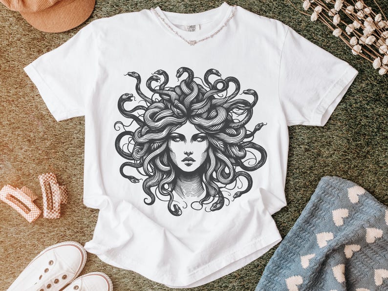 May include: White t-shirt featuring a detailed black and white illustration of Medusa's face, with snakes for hair. The design is centered on the shirt. The shirt is displayed on a textured surface with other accessories.