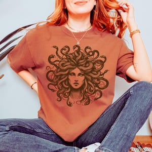 May include: Rust-colored t-shirt featuring a detailed illustration of Medusa's face, with snakes forming her hair. The graphic is in shades of brown. The shirt is a comfortable fit, suitable for casual wear.