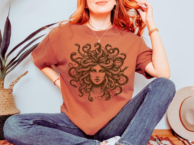 May include: Rust-colored t-shirt featuring a detailed illustration of Medusa's face, with snakes forming her hair. The graphic is in shades of brown. The shirt is a comfortable fit, suitable for casual wear.