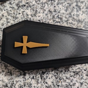 Può includere: Una scatola nera a forma di bara con una croce dorata in cima. La scatola è realizzata in un materiale testurizzato e ha una forma leggermente arrotondata.