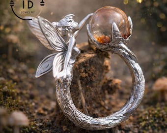 May include: A silver ring featuring a fairy with outstretched wings, holding a clear orb with amber inclusions. The ring's band is textured to resemble tree bark. The ring is set against a natural, mossy background.