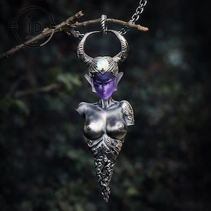 Può includere: Una collana con pendente argentato raffigurante una figura femminile stilizzata con corna, viso viola e orecchie a punta. Il pendente presenta dettagli intricati ed è sospeso a una catena. Il busto della figura è parzialmente scolpito con motivi ornamentali.