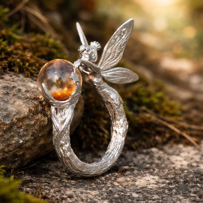 May include: A silver ring depicting a fairy with detailed wings, holding a clear orb with orange and brown inclusions. The ring's band has a textured, organic design. The background shows moss and rock, with a blurred effect. The fairy is the central focus.