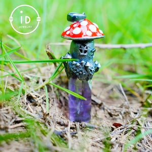 May include: A whimsical mushroom-shaped pendant with a red cap and white spots, atop a detailed, dark-colored structure. The pendant features a purple crystal base and is set against a natural, grassy background. The pendant has the text "iD JEWELRY" in a circle.