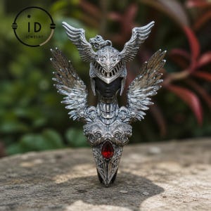 Puede incluir: Anillo plateado detallado con un diseño de guerrero alado. Presenta un rostro con casco, armadura ornamentada y una gema roja central. Las alas están extendidas, sobre un fondo natural. Una pieza de joyería.