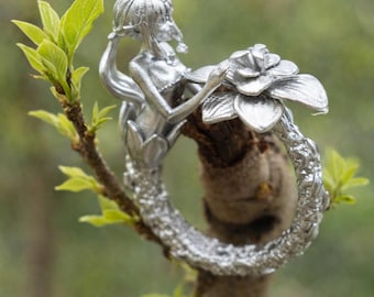 May include: A silver-colored ring featuring a mermaid design intertwined with a flower. The mermaid is sculpted with flowing hair and delicate features. The ring is set against a natural background of a tree branch with green leaves.