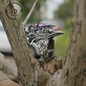 May include: A silver-toned ring featuring a detailed raven design with an amethyst-colored gemstone. The ring is intricately carved, showcasing feathers and a bird's beak. The ring is displayed on a tree branch, highlighting its craftsmanship.