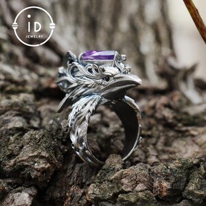 May include: A silver-colored ring with a detailed eagle head design, featuring a purple gemstone. The ring is set against a textured, brown background. The ring has the text "id JEWELRY" in a circular logo.