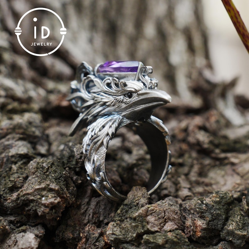 May include: A silver-colored ring with a detailed eagle head design, featuring a purple gemstone. The ring is set against a textured, brown background. The ring has the text "id JEWELRY" in a circular logo.