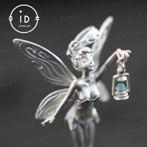 Könnte beinhalten: Silberner Anhänger mit einer Fee, die eine Laterne mit einem türkisfarbenen Edelstein hält. Die Fee hat Flügel und ein fließendes Kleid. Der Anhänger ist ein detailliertes und filigranes Schmuckstück.