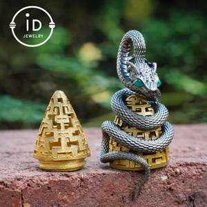 Op de afbeelding: Een gedetailleerde foto van twee decoratieve metalen objecten. Eén is een goudkleurige, opengewerkte kegel. De andere is een zilverkleurige slang die zich rond een goudkleurige basis met groene ogen wikkelt. De afbeelding bevat de tekst "iD JEWELRY" in een witte cirkel.