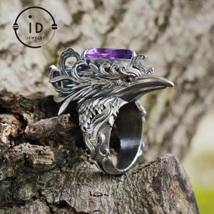 May include: A detailed silver ring featuring a raven design with an amethyst-colored rectangular gemstone. The ring has intricate feather and scrollwork details, with the ID Jewelry logo visible in the background.