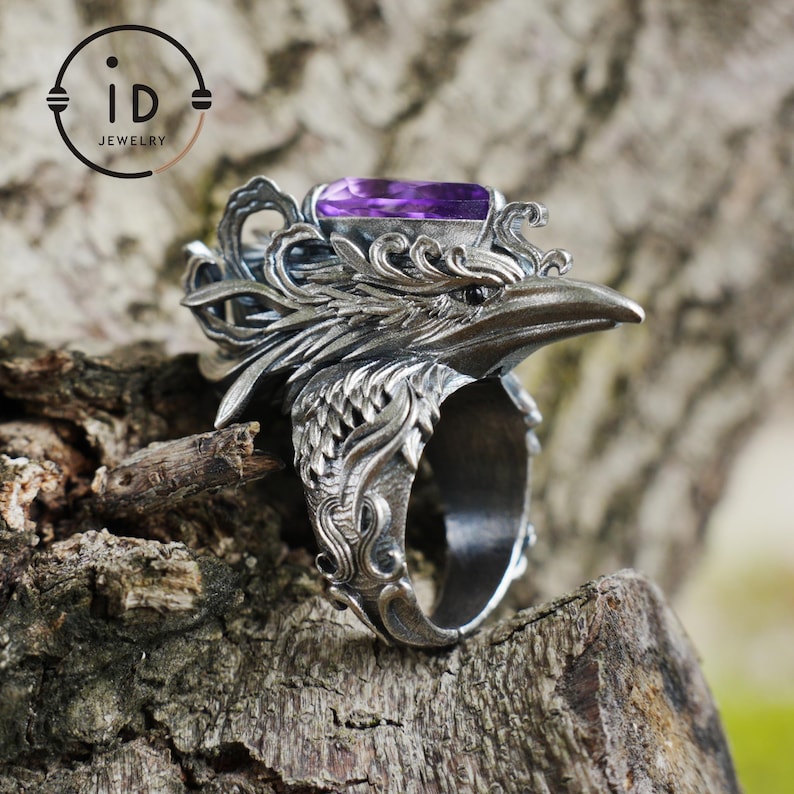 May include: A detailed silver ring featuring a raven design with an amethyst-colored rectangular gemstone. The ring has intricate feather and scrollwork details, with the ID Jewelry logo visible in the background.