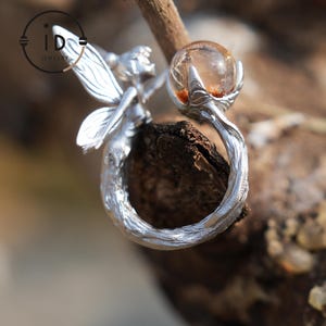May include: A silver ring depicting a fairy cradling a clear orb with amber inclusions. The ring's band is crafted to mimic a tree branch, with detailed wings and body of the fairy. The ring is set against a natural, blurred background.