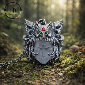 Könnte beinhalten: Eine silberne Anhängerkette mit einem detaillierten, geformten Gesicht mit geschlossenen Augen. Der Anhänger ist mit einem wolfsähnlichen Kopfschmuck verziert, der mit roten und grünen Edelsteinen akzentuiert ist. Die Kette wird auf einer moosigen Oberfläche präsentiert.