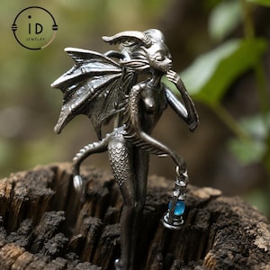 Pode incluir: Estatueta prateada de uma criatura mítica com asas, cauda escamosa e uma lanterna de orbe azul. A figura está pousada sobre uma superfície de madeira desgastada, com o logotipo "id JEWELRY" no canto superior esquerdo. O fundo é folhagem verde desfocada.