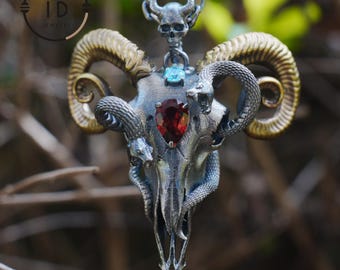 May include: A silver-toned pendant necklace featuring a ram skull design with gold-colored horns. The skull has a red gemstone at the center and is adorned with snakes. A small skull and blue gemstone are at the top. The necklace is suspended from a silver chain.