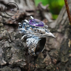 May include: A silver-colored ring with a bird-like design, featuring a purple gemstone. The ring has intricate details, including feathers and a beak. The ring is set against a natural, textured background. The ring is from TD Jewelry.