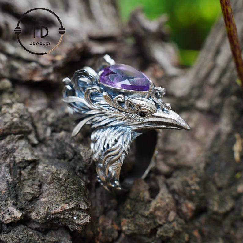 May include: A silver-colored ring with a bird-like design, featuring a purple gemstone. The ring has intricate details, including feathers and a beak. The ring is set against a natural, textured background. The ring is from TD Jewelry.