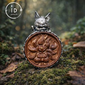 Puede incluir: Un colgante redondo de madera tallada con un diseño de zorro y un marco plateado, coronado por una escultura de zorro plateado. El logotipo de ID Jewelry está en la esquina superior izquierda. El fondo está borroso.
