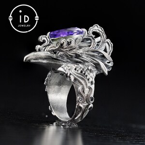 May include: A silver ring featuring an eagle head design with intricate detailing. The ring has a large, rectangular purple gemstone set in the eagle's head. The ring's band is also decorated with ornate patterns. The logo "id JEWELRY" is in the upper left corner.