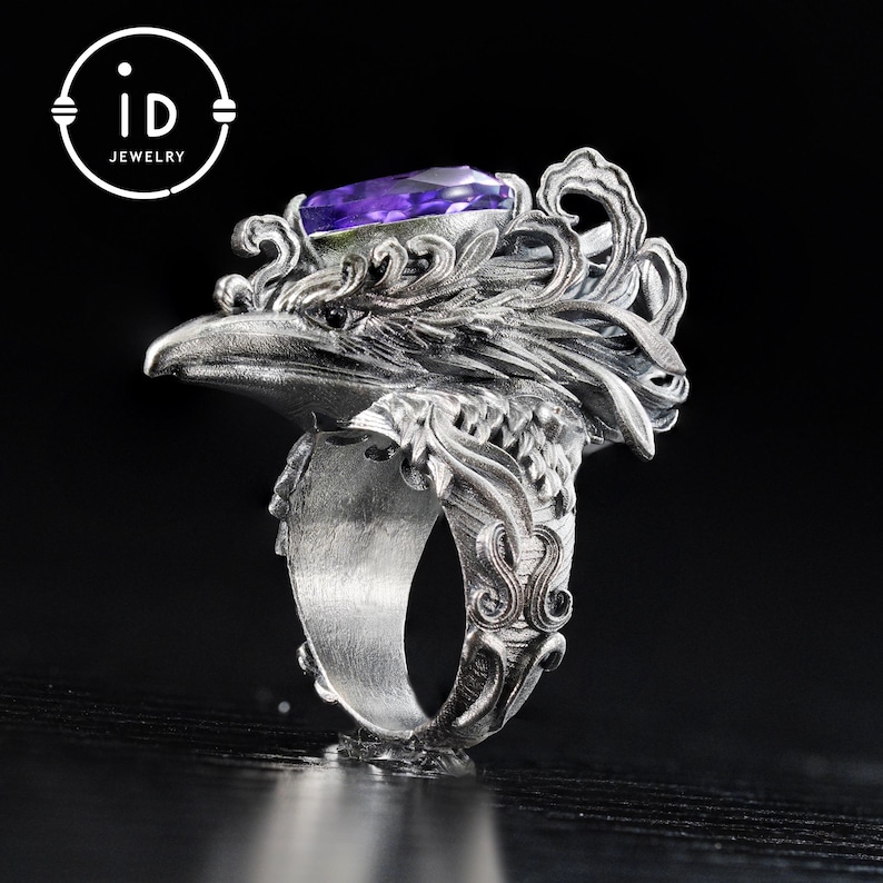 May include: A silver ring featuring an eagle head design with intricate detailing. The ring has a large, rectangular purple gemstone set in the eagle's head. The ring's band is also decorated with ornate patterns. The logo "id JEWELRY" is in the upper left corner.
