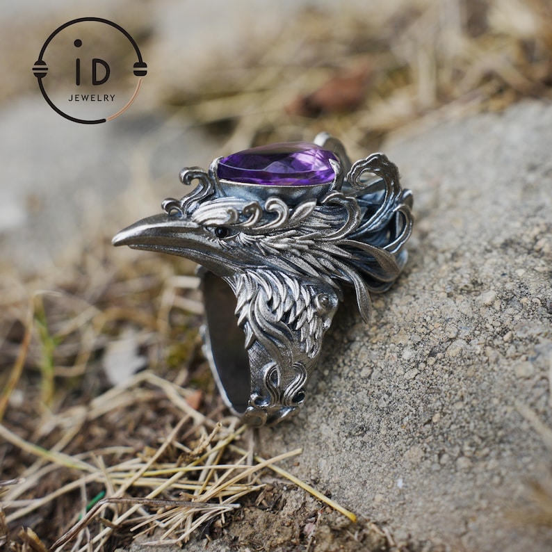May include: A silver ring featuring a sculpted bird head with intricate feather detailing and a pear-shaped purple gemstone set on top. The metal has an oxidized finish, and the logo iD JEWELRY is visible in the upper left corner.