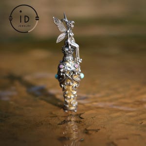 May include: A silver fairy figurine sits atop a gold and silver bottle stopper with pink and turquoise gemstones. The stopper is ornate and detailed, with a swirling design.