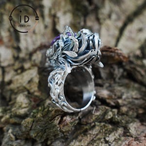 May include: A silver ring featuring an intricate, layered petal design with a dark, oxidized finish and a central purple gemstone. The band is detailed with ornate, carved scrollwork patterns that extend from the base to the top setting.