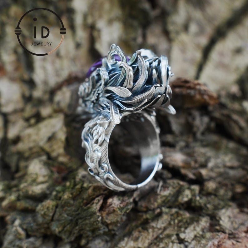 May include: A silver ring featuring an intricate, layered petal design with a dark, oxidized finish and a central purple gemstone. The band is detailed with ornate, carved scrollwork patterns that extend from the base to the top setting.